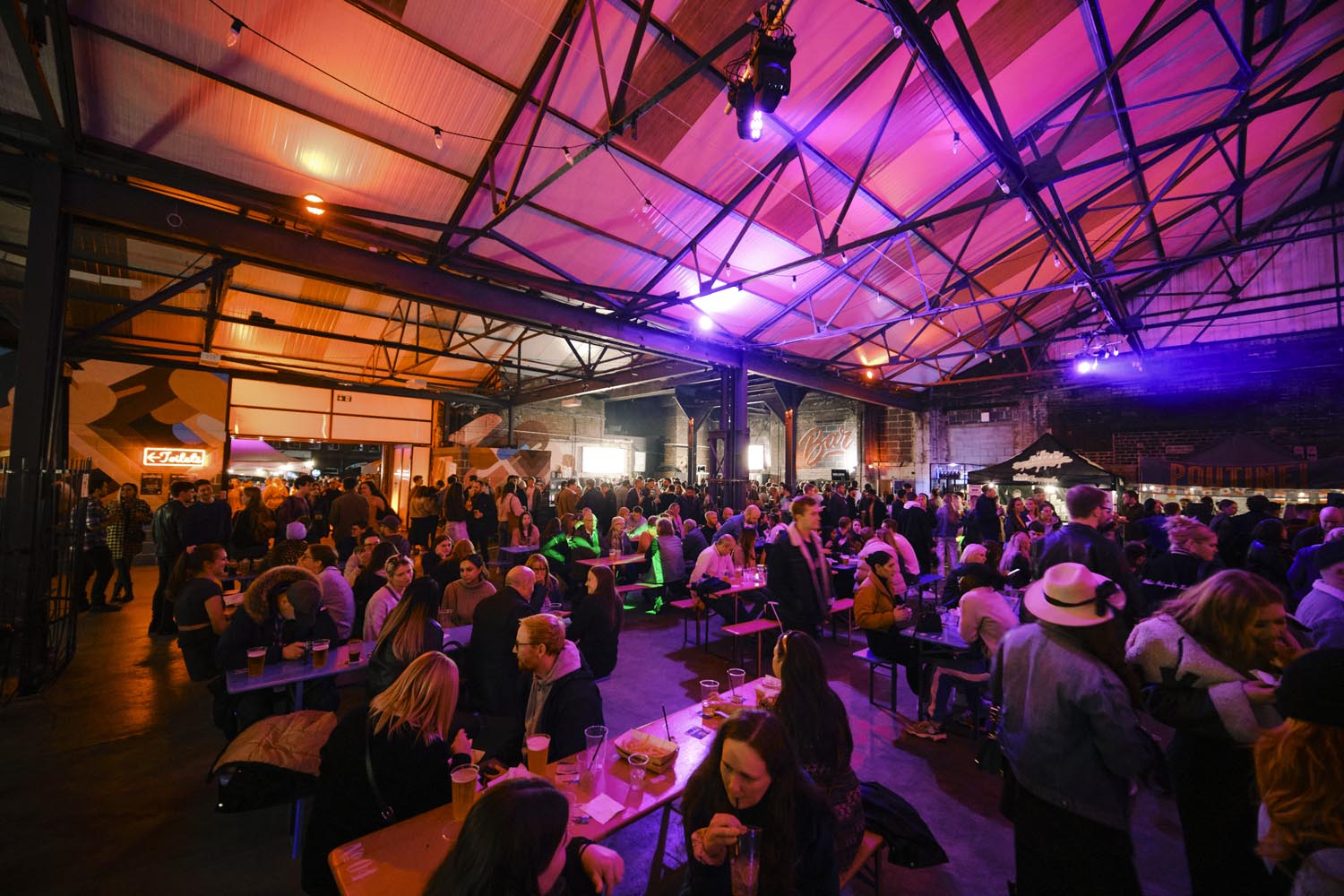 Large indoor venue with high industrial-style ceiling and exposed beams, filled with people seated at long wooden tables and standing in groups. Warm orange and purple lighting creates a lively atmosphere, with food and drinks visible on tables. A bar and food stalls are in the background.