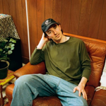 A man in jeans and a green long sleeved t-shirt sits slumped on a brown leather sofa.