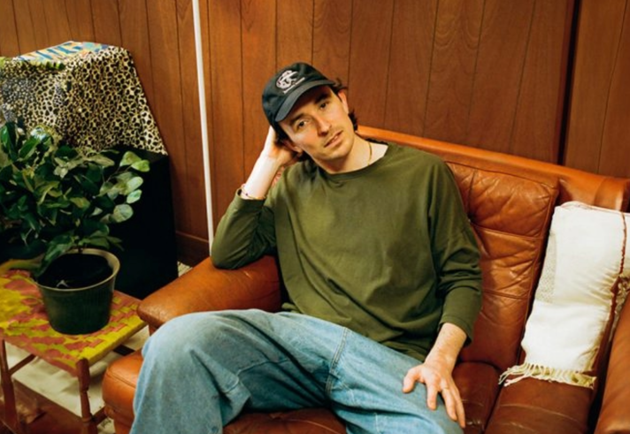A man in jeans and a green long sleeved t-shirt sits slumped on a brown leather sofa.