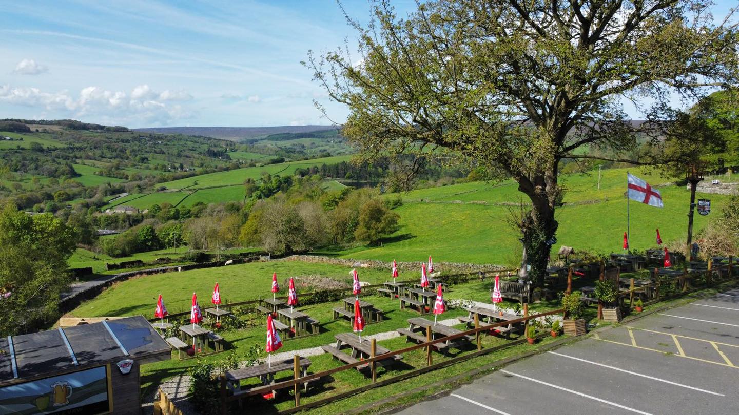 A wide view of The Old Horns Inn beer garden overlooking rolling green countryside, with rows of picnic tables and red parasols set against a backdrop of fields and hills.