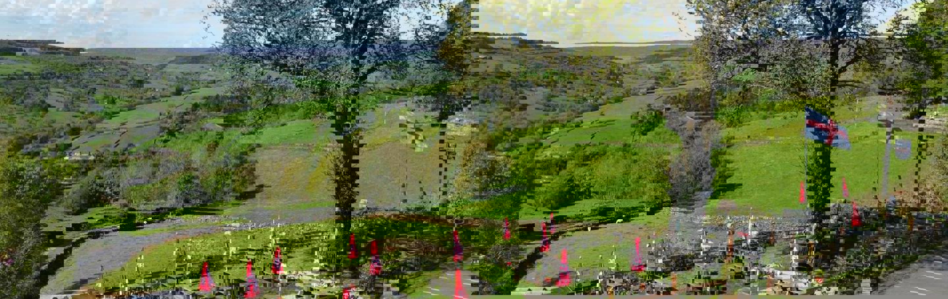 A wide view of The Old Horns Inn beer garden overlooking rolling green countryside, with rows of picnic tables and red parasols set against a backdrop of fields and hills.
