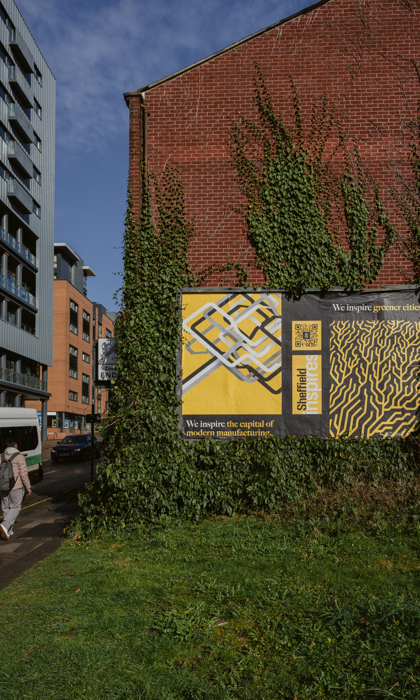 A large brick wall partially covered with green ivy, featuring two posters with bold yellow and black designs. The left poster shows an abstract geometric pattern with the text “We inspire the capital of modern manufacturing.” The right poster has an organic, maze-like pattern with the text “We inspire greater cities.” Both posters include the phrase “Sheffield inspires” and a QR code. In the foreground, there is a grassy area, and to the left, a person walks along the pavement near a white and green bus. 