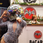 A performer dressed in a sparkling, silver fringed bodysuit poses with one arm raised and the other pointing forward. They wear a large disco‑ball helmet that reflects colourful light. Behind them is a bar area decorated with greenery, branded signs and a circular “Bamboo Door” logo, creating a lively, festive atmosphere.