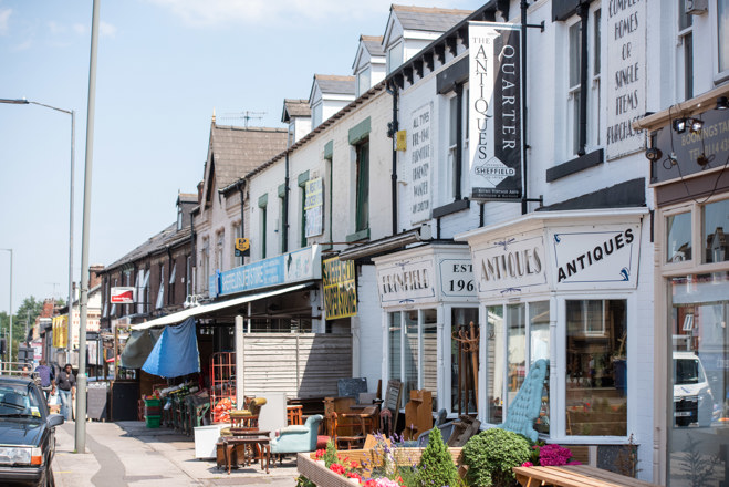 Sunny street scene with independent shops and cafes on both sides, featuring outdoor seating and people walking along the pavement