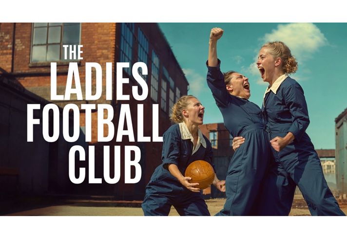 In the courtyard of a factory, three people wearing blue boiler suits scream with joy and triumph. One holds a brown leather ball, one punches the air and one grabs the other's waist. Text reads: THE LADIES FOOTBALL CLUB