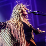 A performer sings passionately into a microphone on stage, arms outstretched, wearing a flowing fringed shawl and layered outfit. Long curly hair catches the stage lights, creating a dramatic effect against a purple-lit background.