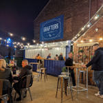 An outdoor bar area at night with string lights overhead, people sitting and standing at high tables, and a blue “Grafters Sheffield” sign mounted on a brick wall above a serving counter.