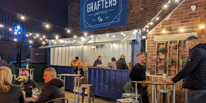 An outdoor bar area at night with string lights overhead, people sitting and standing at high tables, and a blue “Grafters Sheffield” sign mounted on a brick wall above a serving counter.