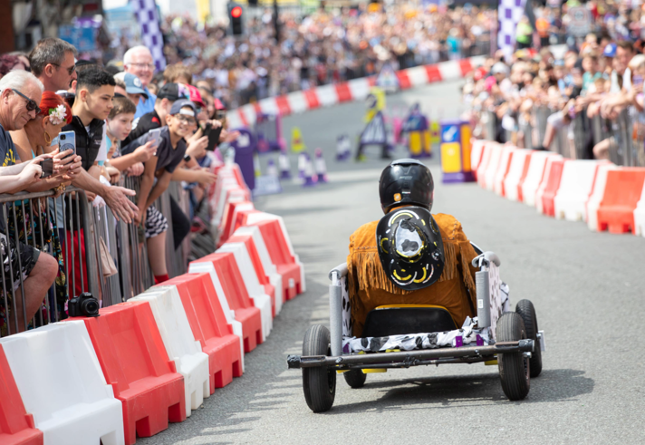 A soapbox cart with a simple frame and four wheels races down a city street lined with red-and-white barriers. The driver wears a black helmet and a fringed brown jacket with a decorative design on the back. Spectators crowd behind metal railings on both sides, watching the event. Purple-and-white race markers and colourful props are visible further down the track.