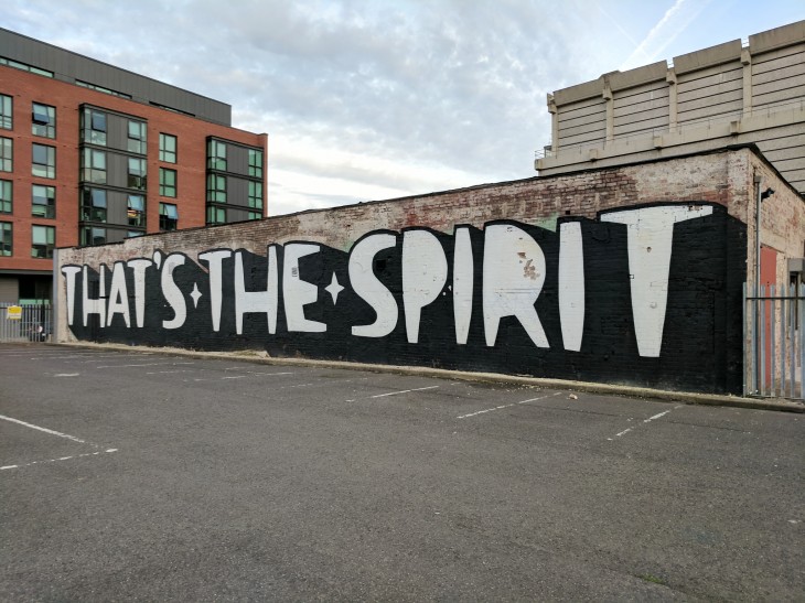 Street art mural on a long brick wall featuring large bold white letters on a black background spelling ‘THAT’S THE SPIRIT’ in an empty parking lot, with modern buildings visible in the background under a cloudy sky.