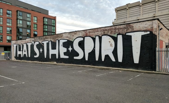 Street art mural on a long brick wall featuring large bold white letters on a black background spelling ‘THAT’S THE SPIRIT’ in an empty parking lot, with modern buildings visible in the background under a cloudy sky.