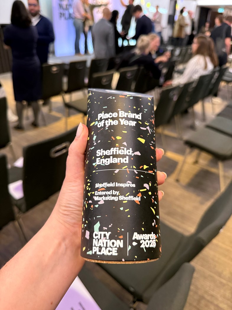 A hand holding a black totem award with white text that reads Place Brand of the Year  with Sheffield, England underneath. In the background members of the conference socialise in front of a presentation stage