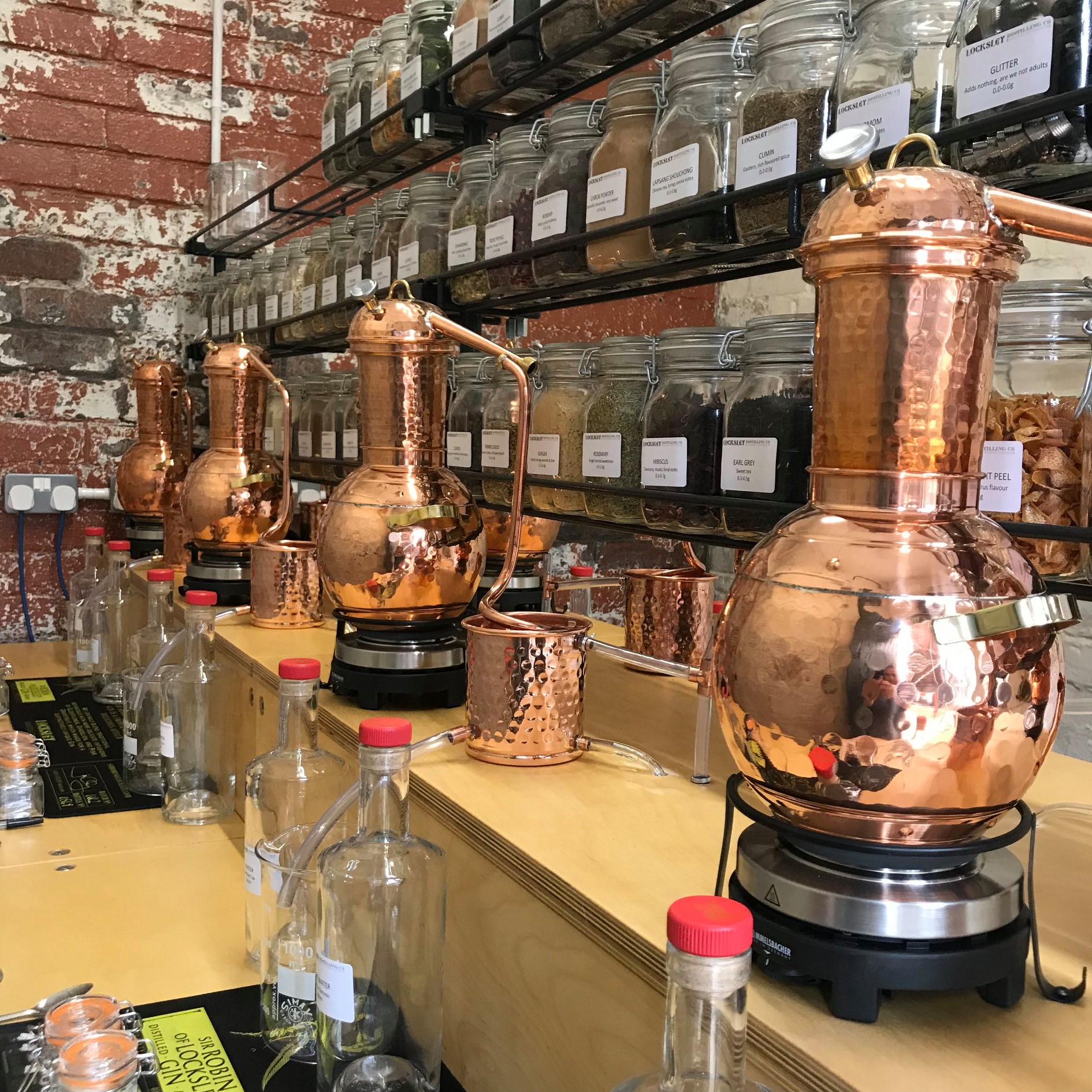 A row of polished copper stills sits on a wooden counter inside a distillery workspace. Shelves above are lined with labelled glass jars filled with herbs, spices, and botanicals. Empty glass bottles with red caps are arranged on the counter, creating an organised, artisanal atmosphere.