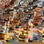 A row of polished copper stills sits on a wooden counter inside a distillery workspace. Shelves above are lined with labelled glass jars filled with herbs, spices, and botanicals. Empty glass bottles with red caps are arranged on the counter, creating an organised, artisanal atmosphere.