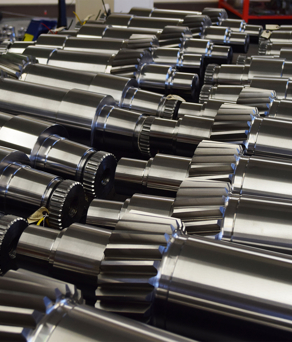 Close-up view of multiple shiny metal gears and cylindrical components arranged in rows. The parts have polished surfaces with visible teeth and grooves, suggesting precision engineering for industrial machinery or mechanical systems.