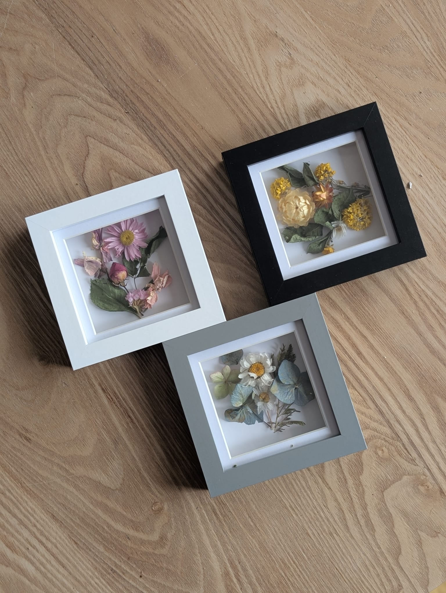 Three square shadow box frames arranged diagonally on a wooden surface. Each frame contains pressed flowers and foliage: one with pink blooms in a white frame, one with yellow flowers in a black frame, and one with mixed greenery and small white flowers in a grey frame.