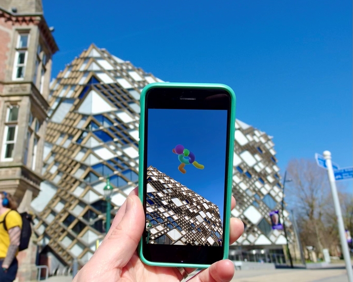 Hand with mobile phone held up in front of The Diamond Building, Sheffield, showing  Look Up AR art 
