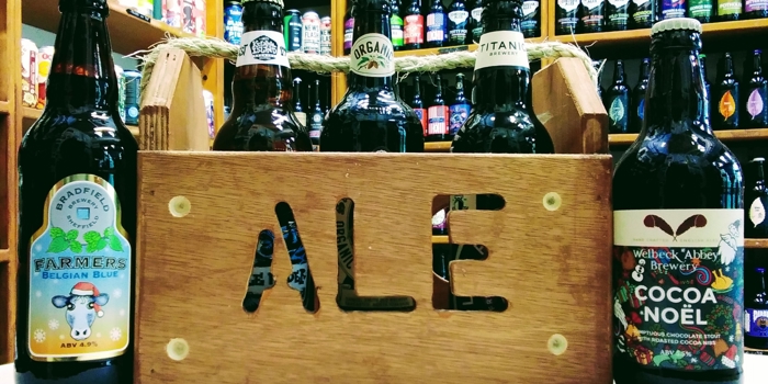 A wooden crate with the word 'Ale' cut into the side, stands on a table. It is filled with bottles of beer, and two more bottles stand on the table.