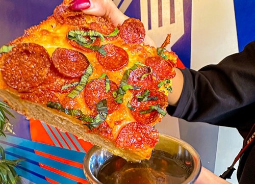 Close-up of a hand holding a large slice of pepperoni pizza topped with fresh basil leaves. The pizza slice is positioned above a metal dipping cup filled with sauce. The background features a colourful wall with bold geometric patterns in blue, red, and white