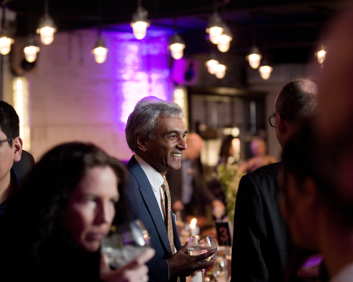 Indoor networking event with people standing and holding drinks in a warmly lit venue. The background features hanging pendant lights, purple accent lighting on the wall, and tables with candles and floral arrangements. A table marker with the number 3 is visible among the decor.
