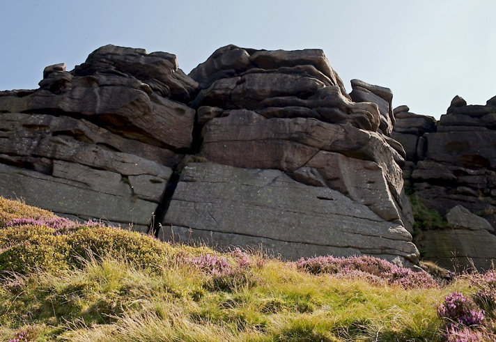 A large rock formation with multiple layers and cracks, situated on a grassy hillside with patches of purple heather. The sky above is clear and bright.
