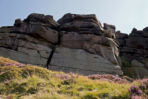 A large rock formation with multiple layers and cracks, situated on a grassy hillside with patches of purple heather. The sky above is clear and bright.