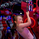 Child wearing a pink and white striped sleeveless top swings on black ropes with red rings in an indoor obstacle course featuring red and gray padded structures and bright overhead lighting.