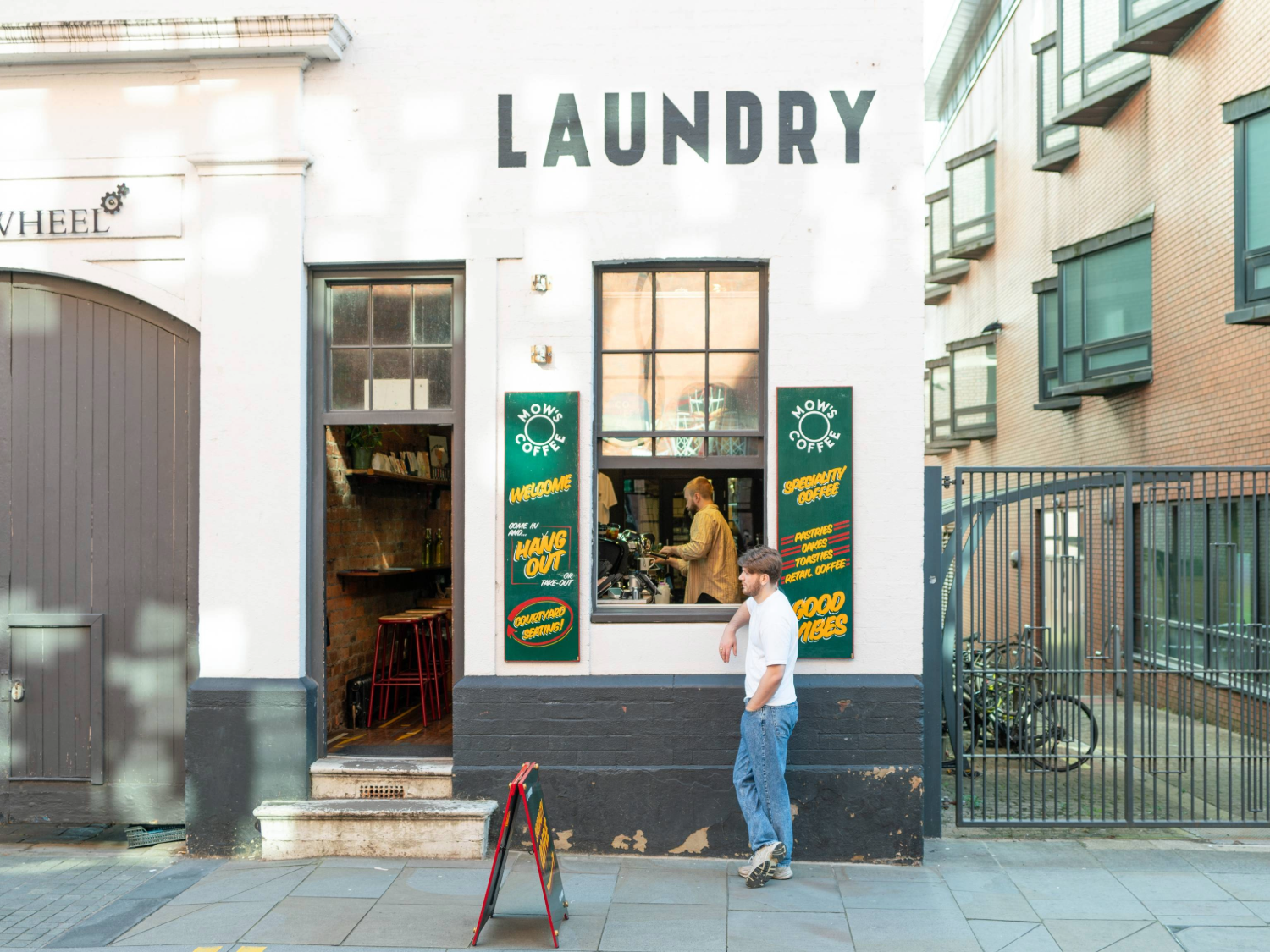 Exterior of a white-painted building with the word “LAUNDRY” in bold black letters above two windows and a doorway. The entrance is open, revealing a rustic interior with bar stools and a counter. Green signs with yellow text advertise coffee and good vibes. A person stands outside on the pavement near a sandwich board, while another is visible inside by the counter. The setting is urban, with a gated area and bicycles to the right.