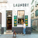 Exterior of a white-painted building with the word “LAUNDRY” in bold black letters above two windows and a doorway. The entrance is open, revealing a rustic interior with bar stools and a counter. Green signs with yellow text advertise coffee and good vibes. A person stands outside on the pavement near a sandwich board, while another is visible inside by the counter. The setting is urban, with a gated area and bicycles to the right.