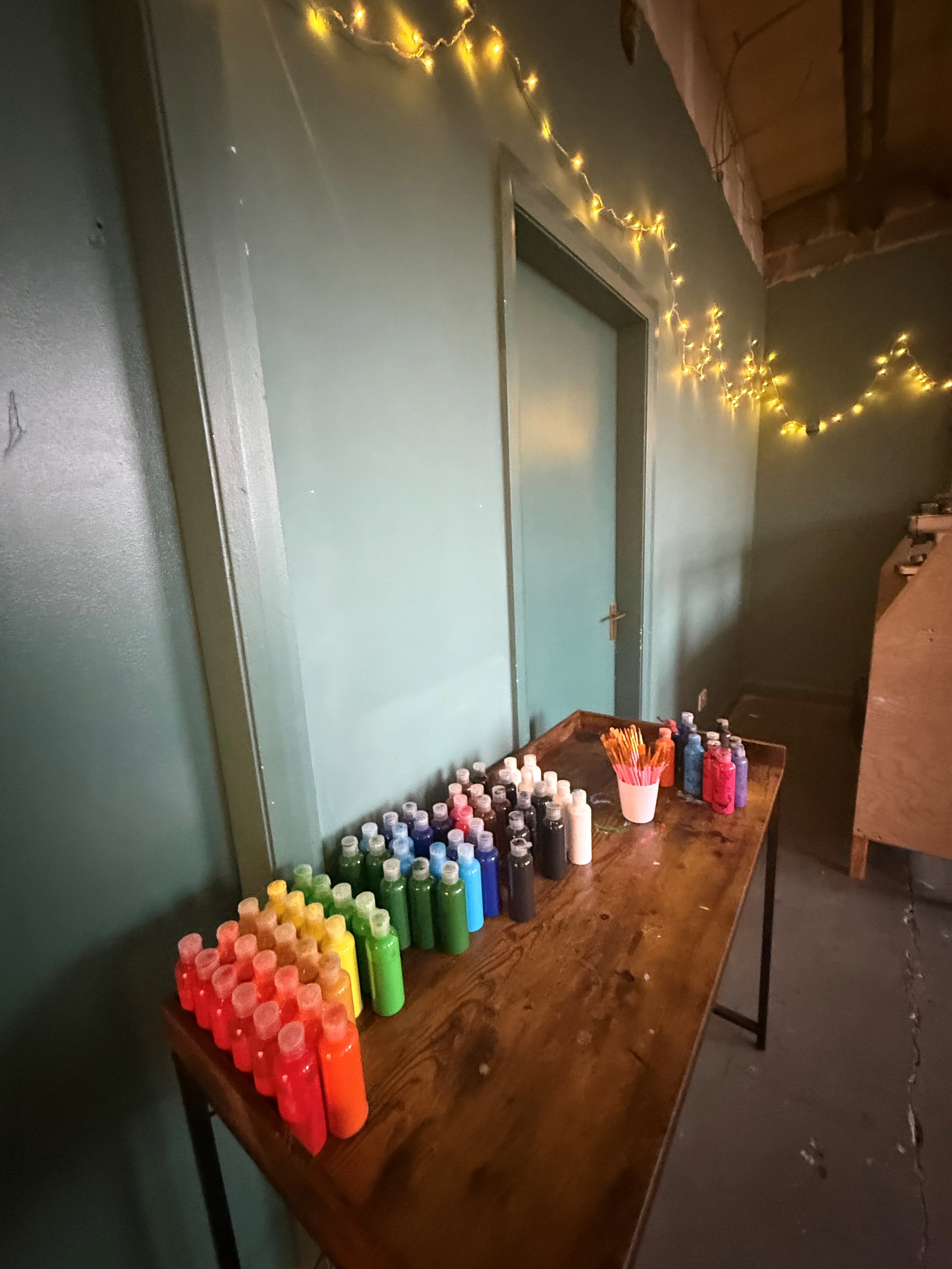 A wooden table lined with rows of brightly coloured paint bottles and pots of brushes, set against a teal wall decorated with warm string lights.