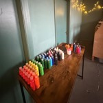 A wooden table lined with rows of brightly coloured paint bottles and pots of brushes, set against a teal wall decorated with warm string lights.