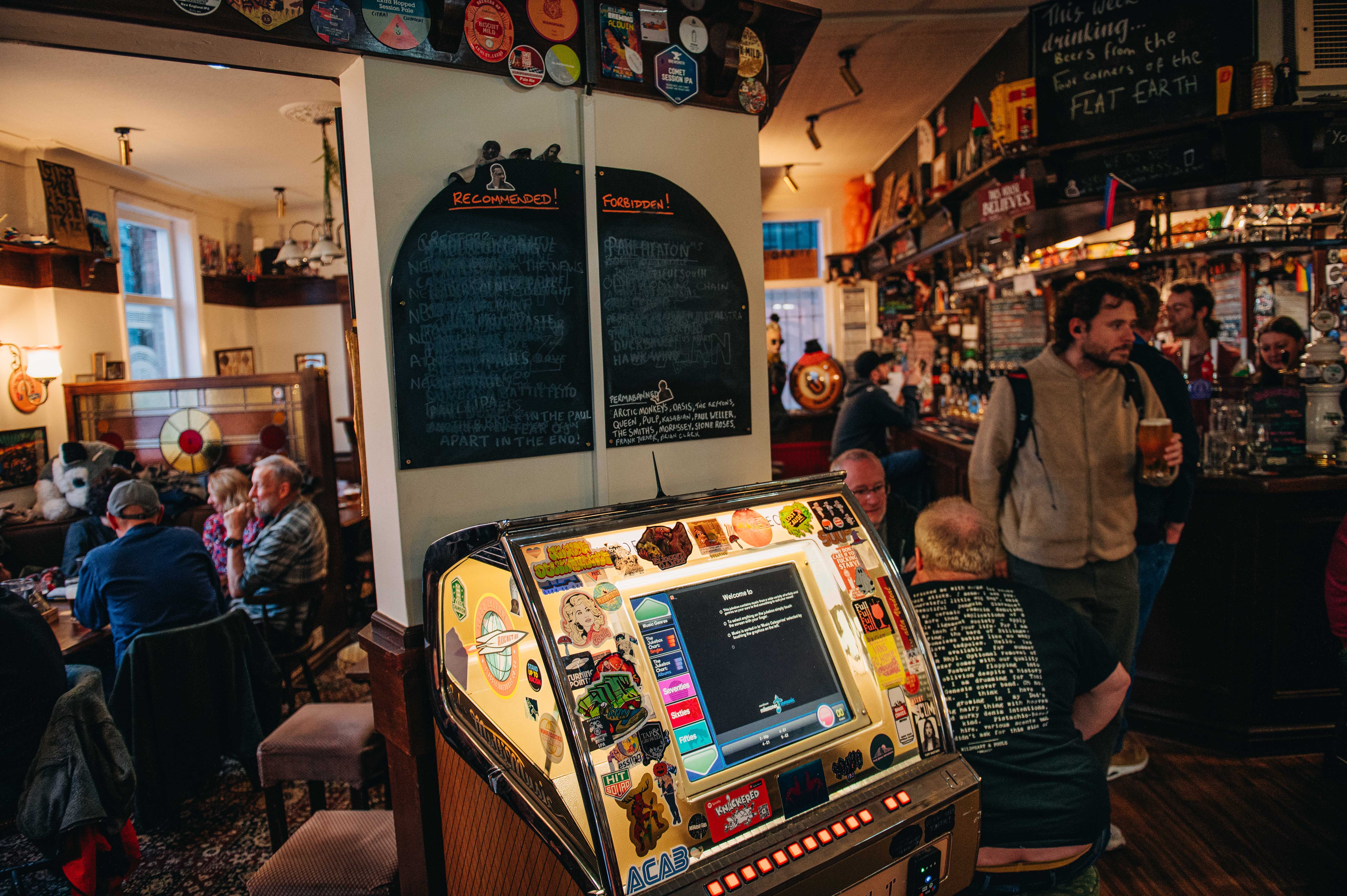 Rutland Arms jukebox lit up, with a chalk board above that's separated into forbidden and recommended. Groups of people are sat to the left of the jukebox, with the bar to the right. 