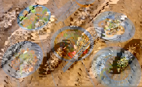 Overhead view of an assortment of Asian-inspired dishes arranged on a wooden table. The spread includes a steamed bun topped with sliced green onions, a plate of crispy fried rings with dipping sauce, a bowl of glazed meat garnished with sesame seeds, a dish of tofu cubes with herbs, and a plate of dumplings in a creamy sauce sprinkled with sesame seeds and sliced chili. Black chopsticks rest on two plates, and a cocktail in a coupe glass is placed near the center.