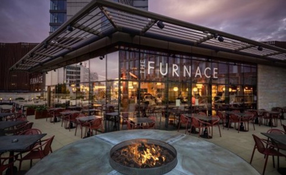 Exterior view of The Furnace restaurant at dusk, featuring a modern glass-fronted building with warm interior lighting. The outdoor terrace is furnished with red chairs and tables, and a large circular fire pit with flames is in the foreground. The sky has a purple hue, and tall buildings are visible in the background.