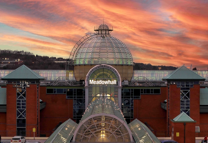 The Meadowhall exterior at sunset, featuring the glass-domed main entrance, red-brick walls and a dramatic orange and pink sky.