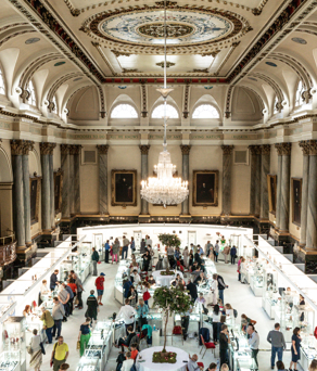 Elegant exhibition inside a grand hall with ornate columns, arched windows, and a decorative ceiling featuring a large chandelier. Visitors browse display booths arranged in a square layout around a central tree.