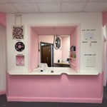  A pink-and-white reception area with a serving hatch, surrounded by signs for various craft activities such as tote bags, vinyl art, air-dry clay and textured art.