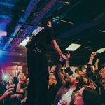 A performer stands on a barrier inside a dark, atmospheric venue, reaching out to audience members who have their hands raised. The room is lit with stage lighting in pinks and blues, and the crowd fills the foreground.