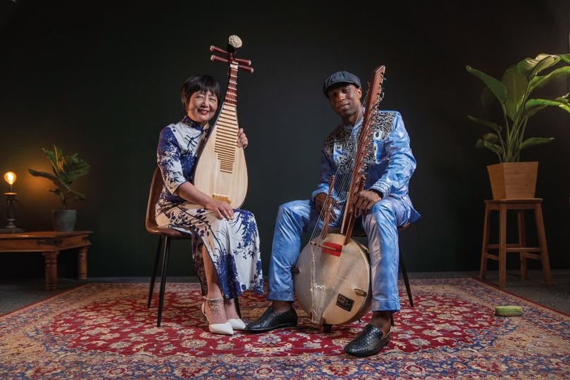 Two people are sat on chairs, smiling at the camera. One is holding a Chinese pipa, and the other is holding a West African kora. There is subdued lighting and behind them, to one side, is a pot plant.