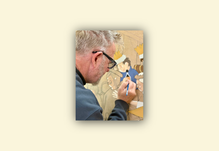 A person is painting a detailed illustration on a textured surface. The artwork features cartoon-style characters wearing festive crowns and fur-trimmed outfits, gathered around a table. One character is holding a bottle, and the scene suggests a celebratory or holiday setting. The painter’s hand is visible holding a fine brush, adding colour to the design with precision.