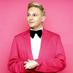 A photo of the performer, who is wearing a pink suit, with a bowtie. They are looking away from the camera. They are stood infront of a pink background