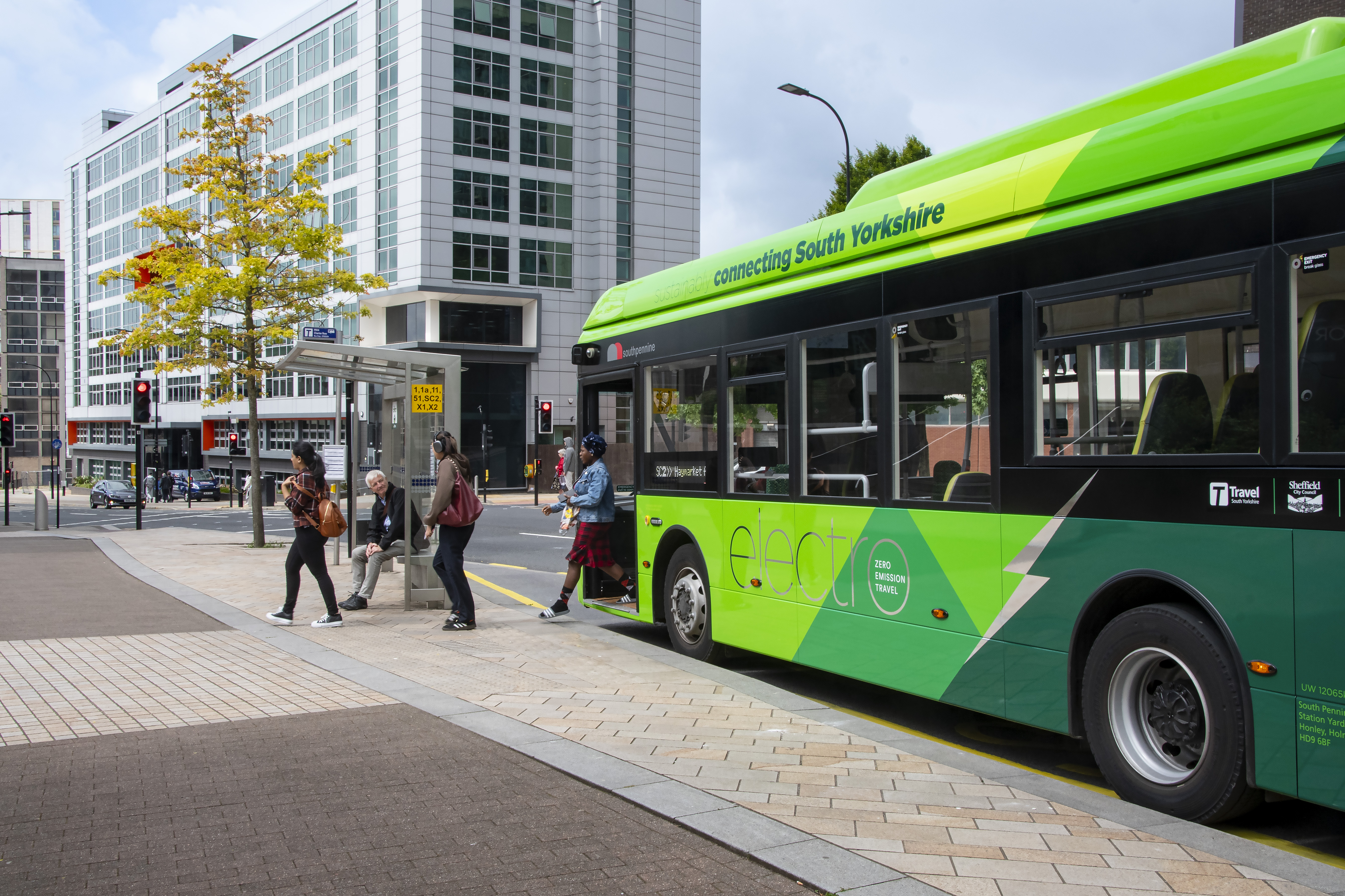 A bright green electric bus with the text “connecting South Yorkshire” on the side is stopped at a bus stop in an urban area. Several people are boarding the bus from a paved sidewalk. The background features modern multi-story office buildings with large glass windows, street signs, and a tree with yellow leaves under a partly cloudy sky.