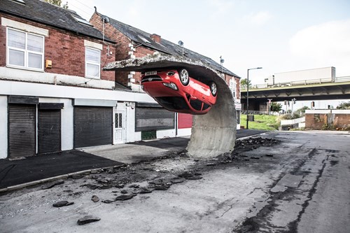 Red car embedded upside down in a large curved concrete structure on a deserted street, with terraced brick buildings and closed shopfronts in the background.
