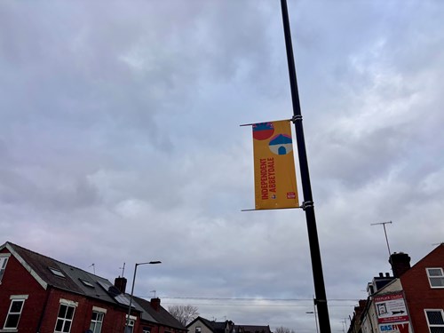 Street banner attached to a black lamppost with text reading ‘Independent and Proud’ in red on a yellow background, featuring colorful abstract shapes. The banner is set against a cloudy sky with rooftops of red-brick houses visible below.